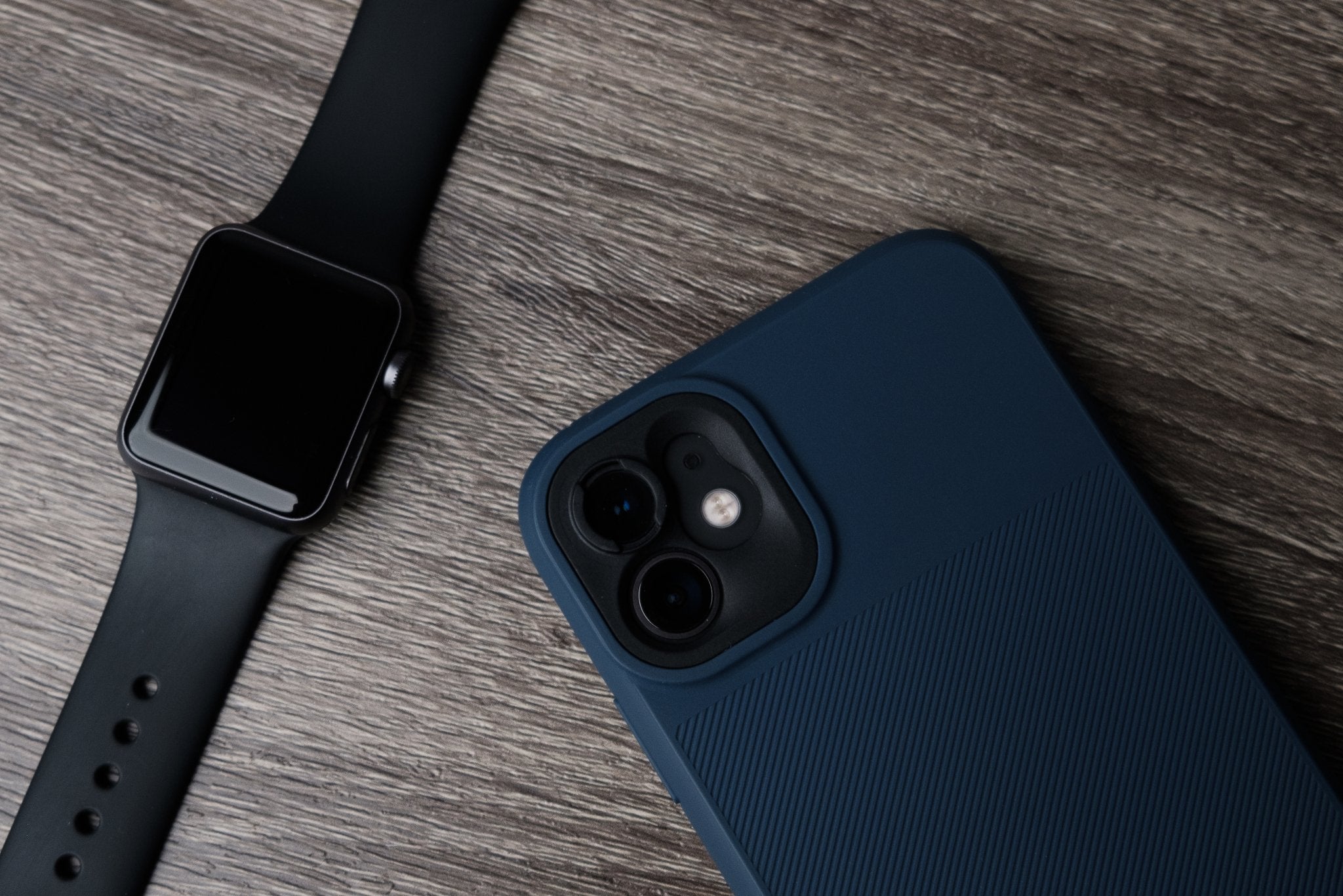 This image shows a close-up view of a black smartwatch and a smartphone with a blue protective case. Both devices are placed on a textured wooden surface, highlighting their sleek designs and tech-focused style.